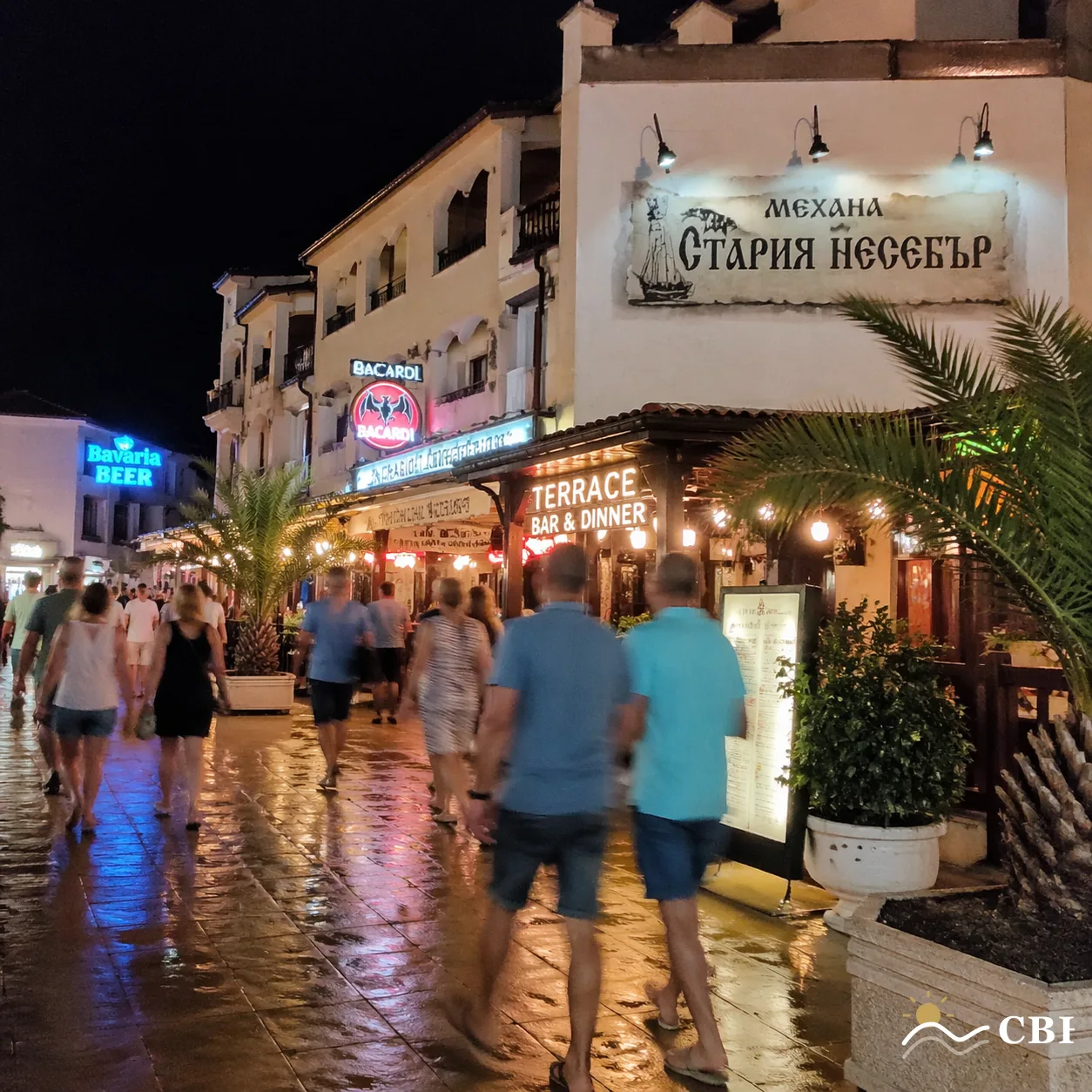 Candid night-time photo of a Sunny Beach tavern strip in Bulgaria with Cyrillic and Latin bar signage, palm trees, wet pavement reflections and blurred holidaymakers walking between bars.