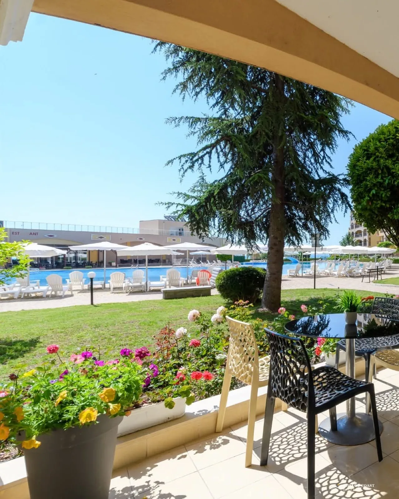View from the Midia reception terrace to the second pool, with flower beds, cedar tree shade, and the restaurant building beyond